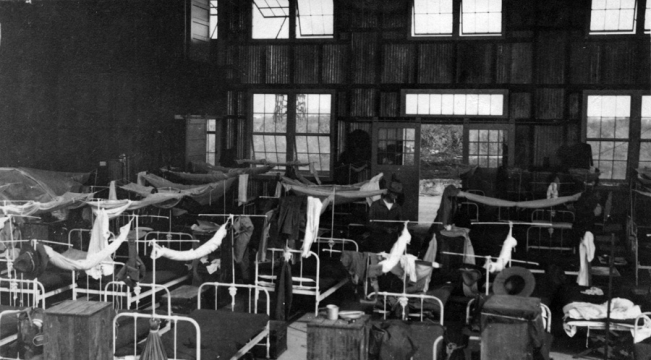 Long building with two rows of windows that has been converted into a barracks. Interior of the barracks showing beds and mosquito netting.  Same building as seen in 2006.040.123