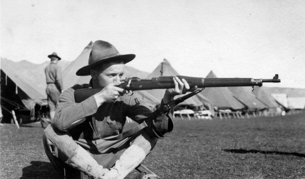 Military man,  sitting on the ground with his legs crossed,  shooting a rifle.  A row of tents in the background.