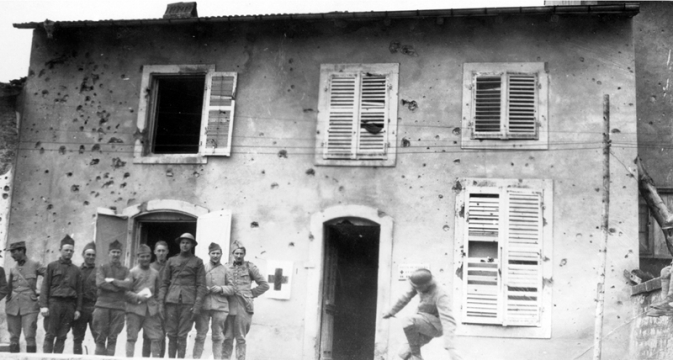 WWI soldiers standing in front of two story building that is being used as a Red Cross Hospital. The building shows signs of being hit with gunfire.